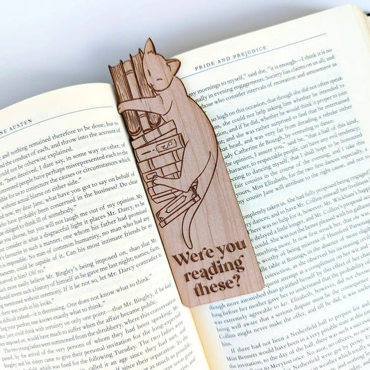 A wooden bookmark of a cat laying on a stack of books that says "were you reading these?"