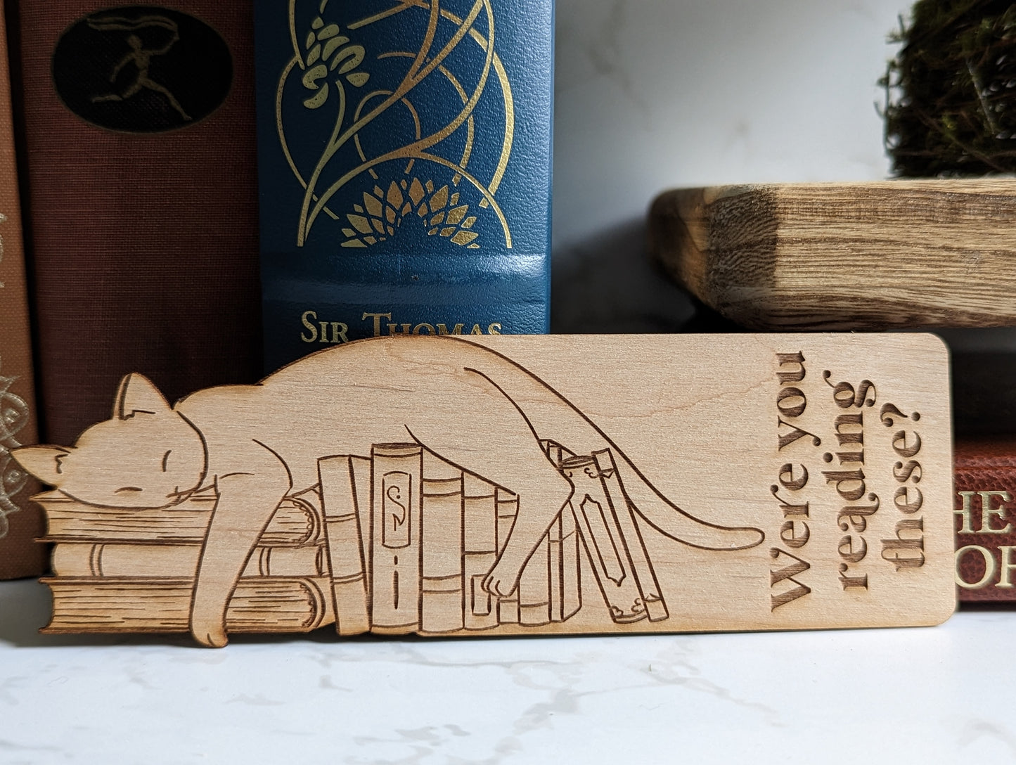 A wooden bookmark of a cat laying on a stack of books that says "were you reading these?"